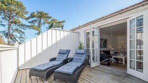 Une terrasse avec deux chaises longues et une table, un mur extérieur blanc et une porte donnant accès à un espace intérieur.