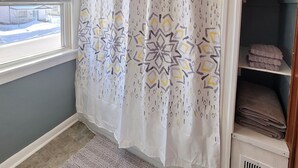 A bathroom with a shower curtain featuring a geometric pattern, a window with blinds, a radiator, and a towel rack with folded towels. - Cute & cozy 1 bedroom apartment - sleeps 4 (Negaunee)