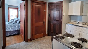 A compact kitchen with a stove and cabinets, a wooden door, and a bedroom visible through an open door. - Cute & cozy 1 bedroom apartment - sleeps 4 (Negaunee)