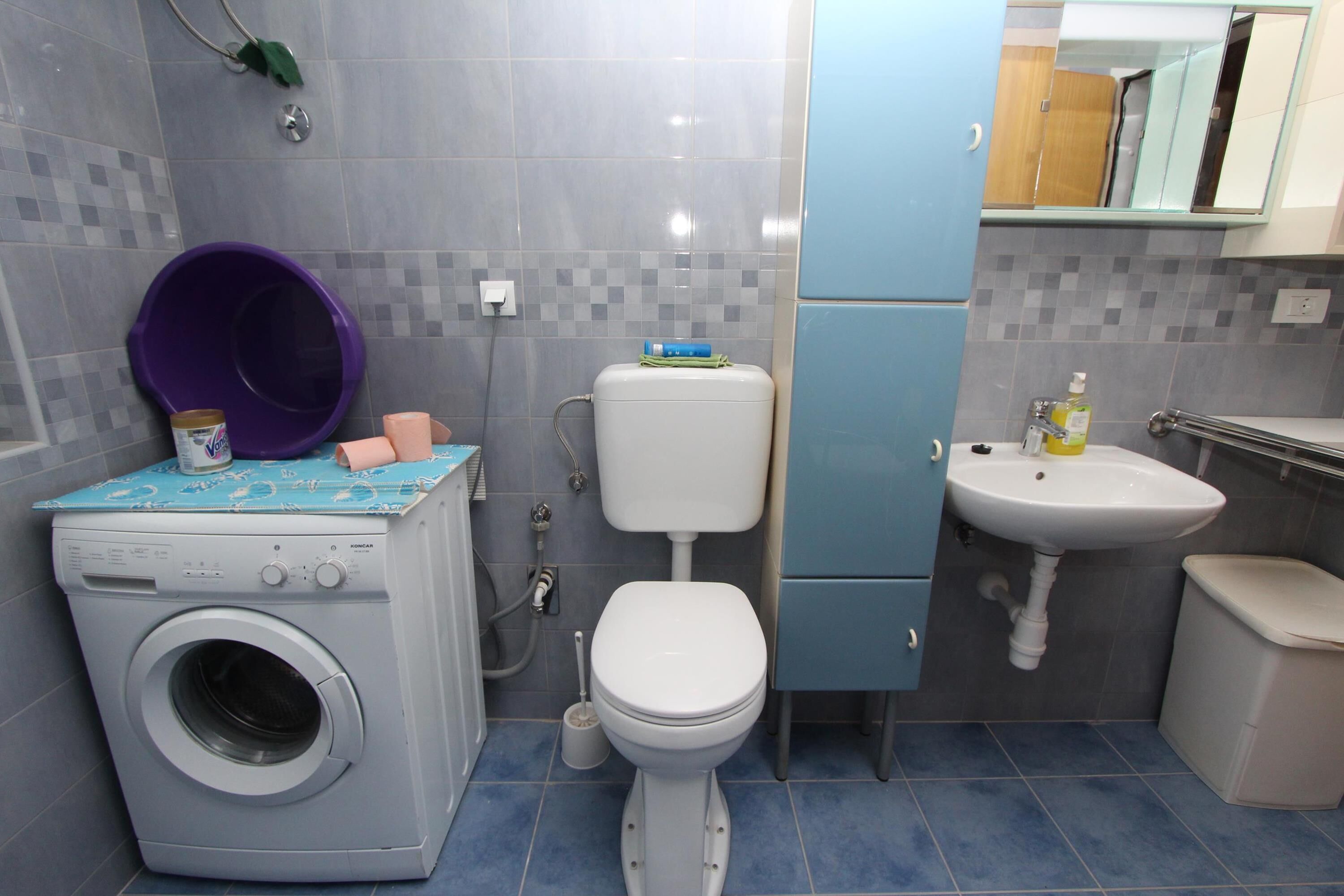 A compact bathroom with a washing machine, toilet, sink, and a small trash bin.