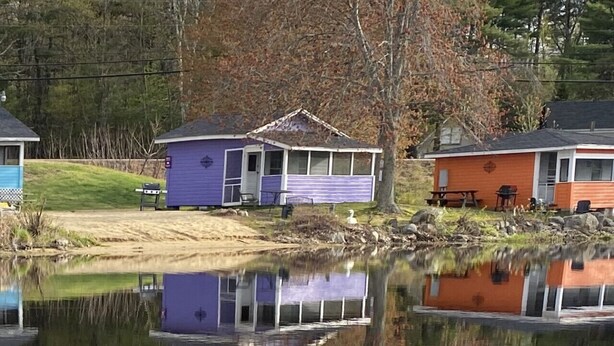 A lakeside cabin with a reflection in the water, surrounded by trees and a grassy area. - Lakefront Purple Cottage (Northwood)
