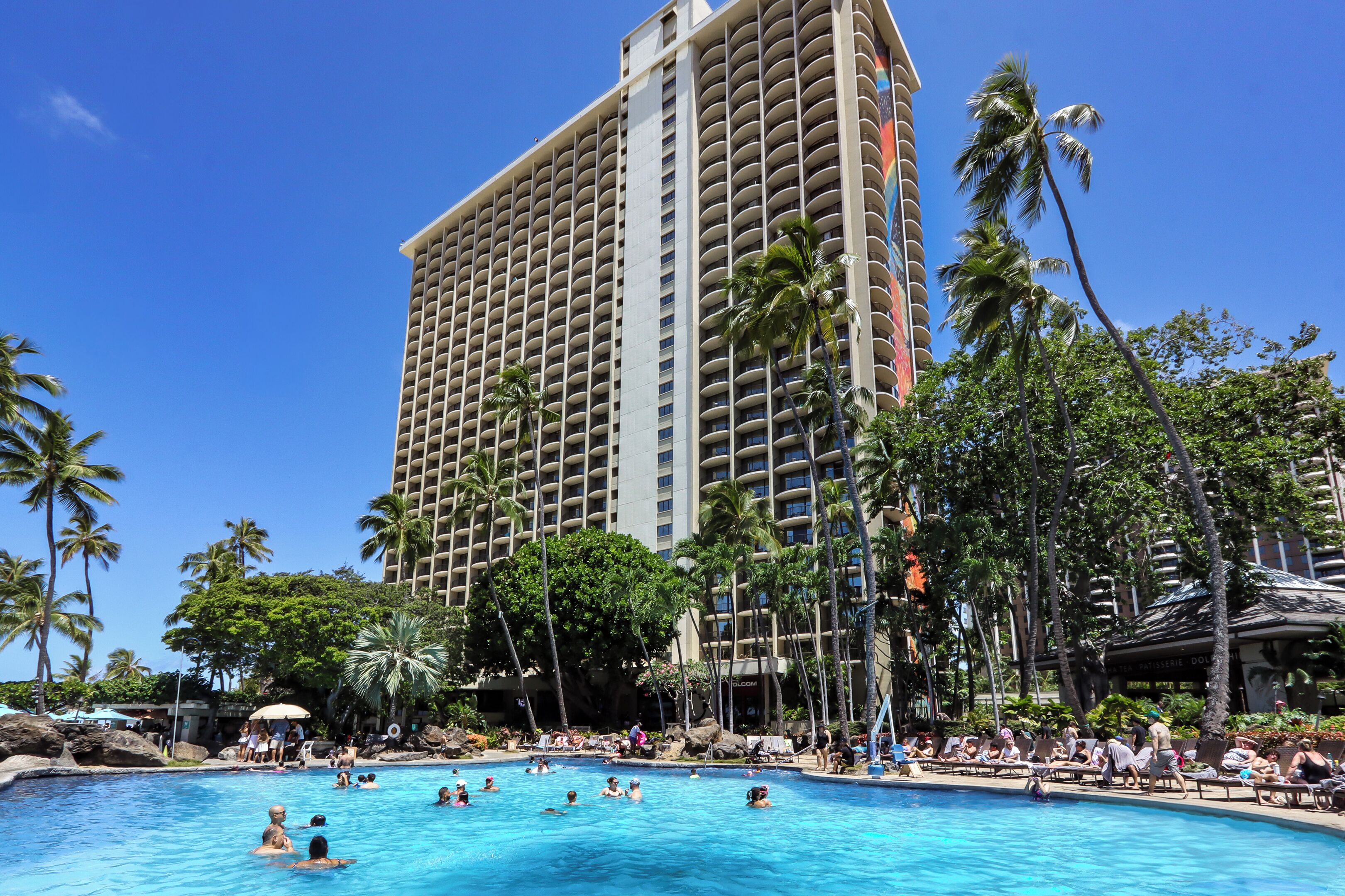 Un hôtel doté d’une grande piscine entourée de palmiers, avec un immeuble imposant en arrière-plan.