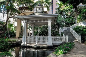 A gazebo with a white railing, a small pond, and a staircase leading up to a building.