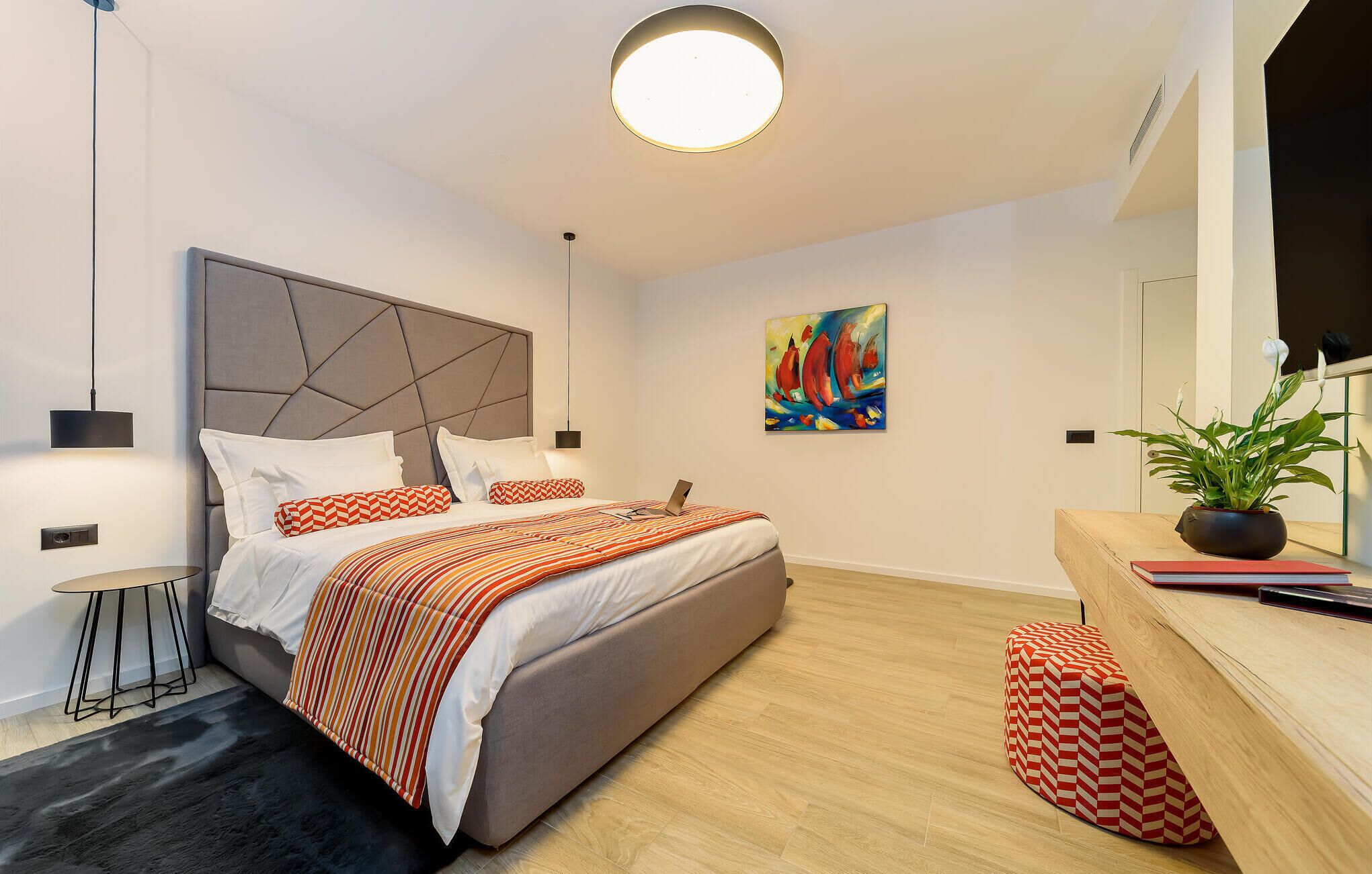 A modern bedroom with a large bed, a bedside table, a small side table, a potted plant, and a painting on the wall.