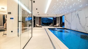 A modern indoor swimming pool with glass walls, lounge chairs, and a view of a mountainous landscape. - Luxurious home in Marina with sauna (Marina)