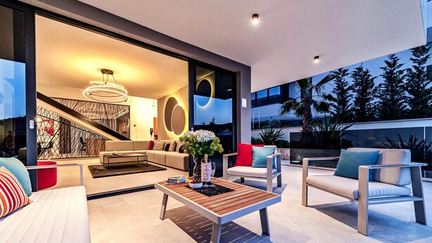 A modern living room with a sofa, coffee table, and chairs, a staircase, and a large window with a view of trees. - Luxurious home in Marina with sauna (Marina)