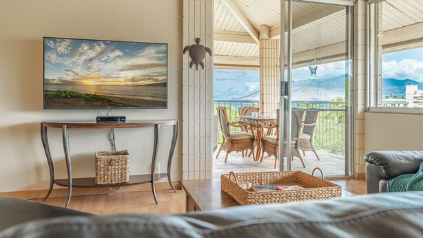 Een moderne woonkamer met een grote televisie, een houten tafel, rieten manden en een balkon met riemeisen meubilair.