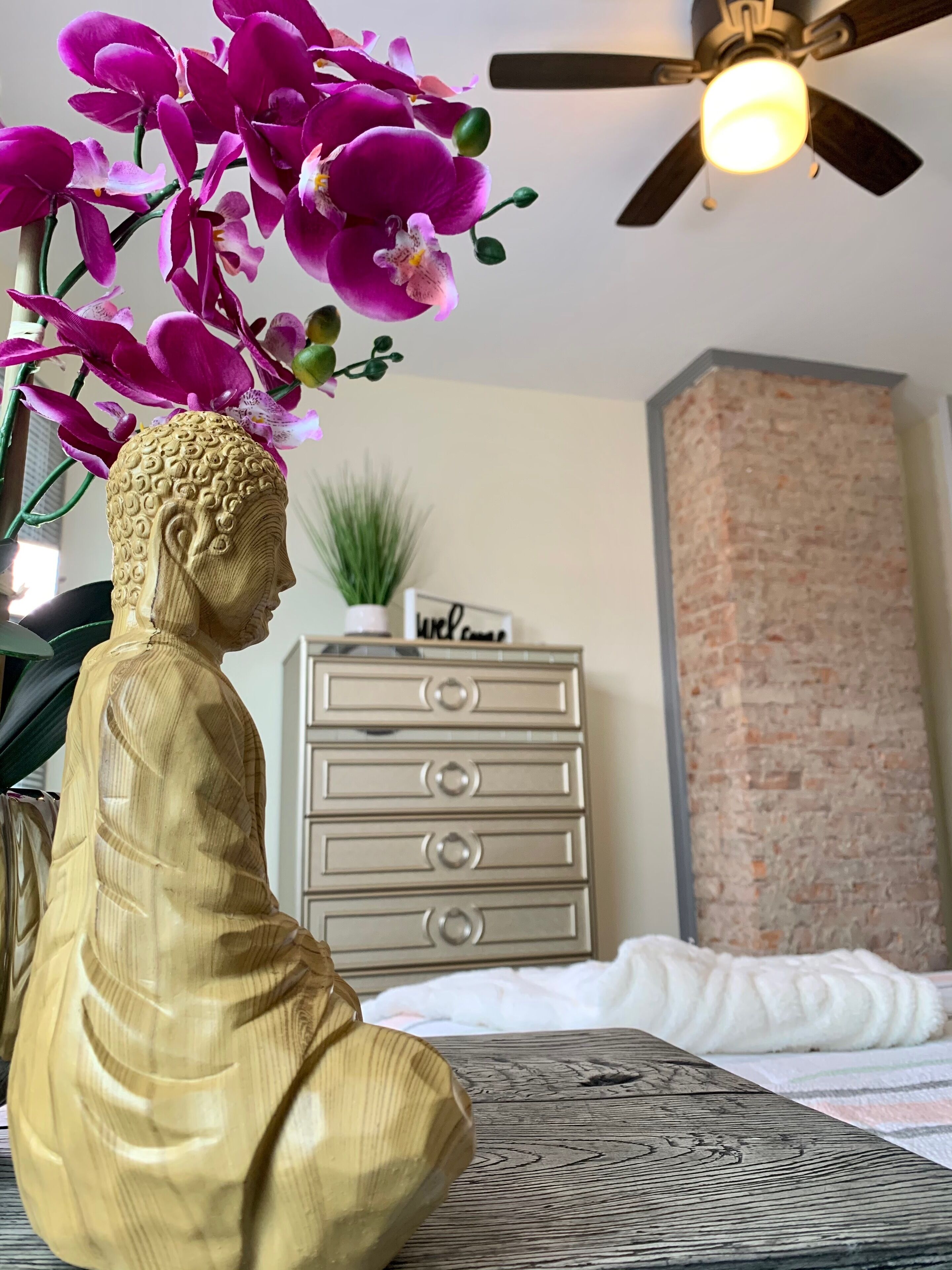 A Buddha statue, a ceiling fan, a brick wall, a dresser, and a sign with "Welcome" on it.