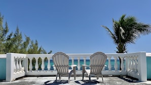 Two chairs and a small table on a balcony with a white balustrade, overlooking a clear blue sea and palm trees. - Quaint Studio at Belizean Reef Suites, Short Walk to Everything! (San Pedro - Town)