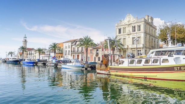 Un port de plaisance avec des bateaux amarrés, un bâtiment imposant à l’architecture classique et un phare en arrière-plan.