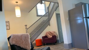 A modern living room with a black leather chair, a fluffy throw, and a red ottoman. A staircase with a metal railing leads to a second floor. A large gray refrigerator is visible on the right.