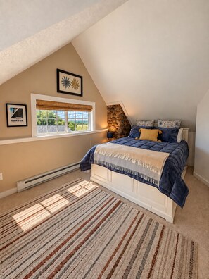 A bedroom with a sloped ceiling, a bed with a blue duvet, a stone wall, a window with a view, and a radiator.