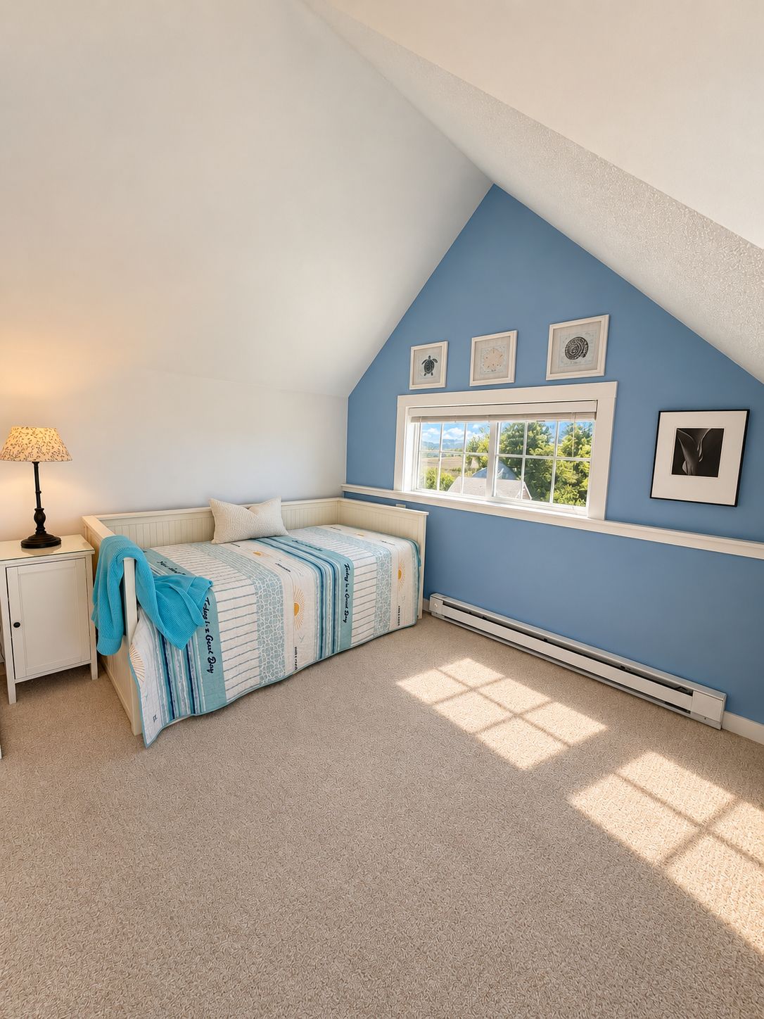 A bedroom with a sloped ceiling, a bed with a striped bedspread, a nightstand with a lamp, a window with a view, and wall-mounted artwork.