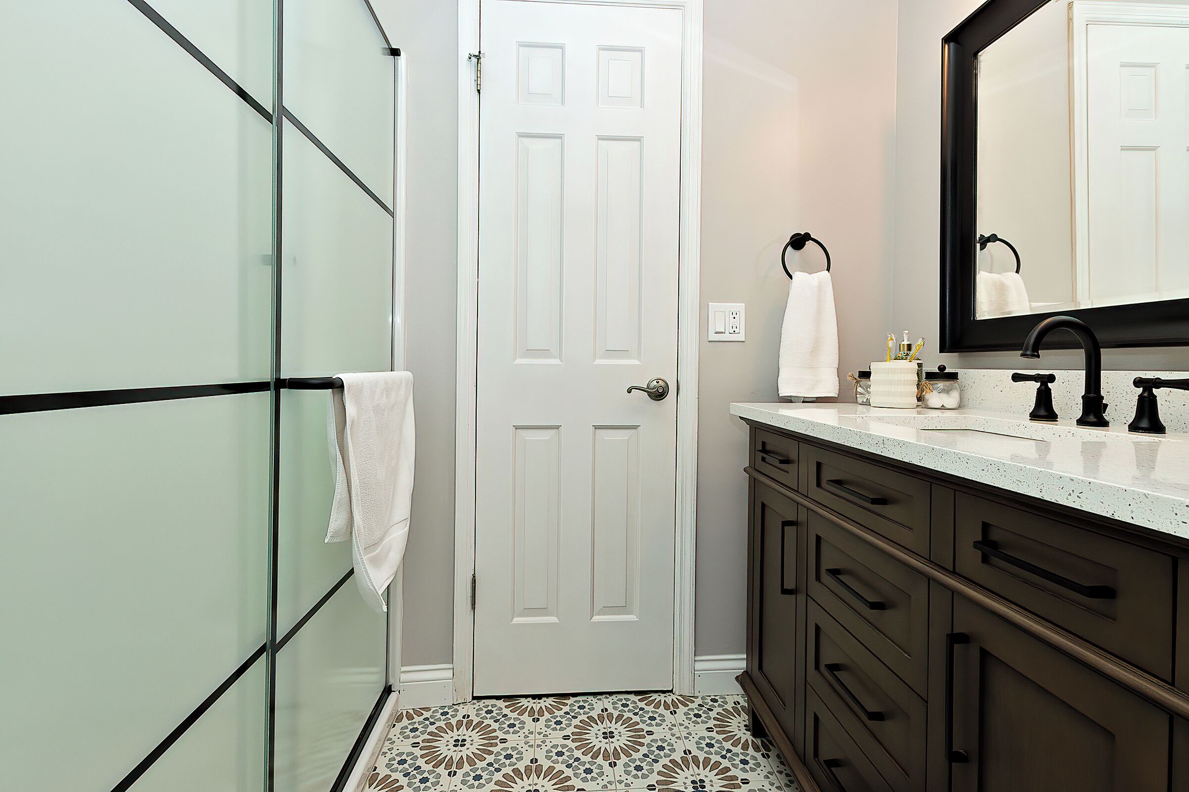 A bathroom with a white door, a glass shower enclosure, a sink with a black faucet, and a tiled floor.