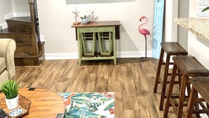 Una sala de estar con piso de madera, un televisor montado en la pared, un aparador verde, un banco de madera y un tapete colorido.