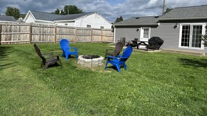 Ein Garten mit Feuerstelle, zwei blauen Adirondack-Stühlen, einem Holztisch und einem Grill.