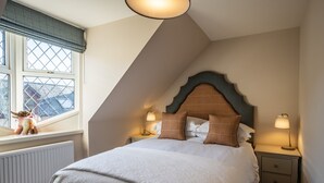 A bedroom with a sloped ceiling, a bed with white bedding, two bedside lamps, and a window with a view of buildings. - Contemporary Cottage in Ballater Village (Ballater)