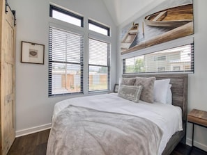 A bedroom with a large bed, two windows with blinds, a wooden headboard, and a wall-mounted artwork of boats. - The Heather @ Water 65, Tiny Home walk to downtown (Waynesville)