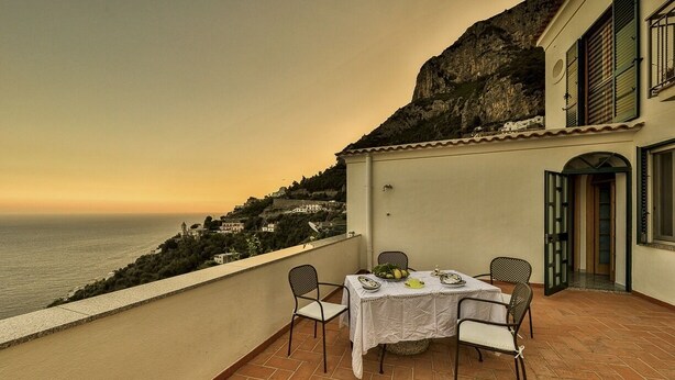 Eine Terrasse mit Tisch für zwei Personen mit Blick auf die Küste und Gebäude an einem Hügel.