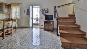 Ein Wohnzimmer mit Fliesenboden, einer Holztreppe, einem Fernseher und einem Essbereich mit Tisch und Stühlen.