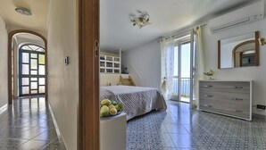 Ein Schlafzimmer mit einem Bett, einem Schminktisch und einem kleinen Tisch mit Obst. Ein Fenster mit Vorhängen und eine Tür zu einem anderen Raum.
