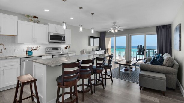 A modern kitchen with white cabinets, a central island, and bar stools. There is a dining area with a table and chairs, and a balcony with a view of the ocean. - REMODELED! BeachFront, Bch Service, Pool, wifi, Kitchen, grill (Destin)