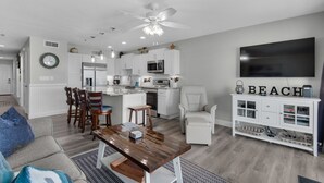 A modern kitchen with white cabinets, a built-in oven, and a microwave. There is a dining area with a wooden table and chairs, and a living room with a white sofa and a TV mounted on the wall. - REMODELED! BeachFront, Bch Service, Pool, wifi, Kitchen, grill (Destin)