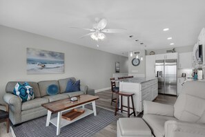 A modern living room with a gray sofa, a wooden coffee table, and a white armchair. There is a kitchen area with white cabinets and a stainless steel refrigerator. A ceiling fan and pendant lights are visible above the dining table.