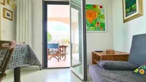 A living room with a sofa, a table, and a chair. There is a painting on the wall and a window with a view of the outside.