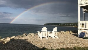 A beach with white chairs and a deck overlooking the ocean, a rainbow, and a rocky shoreline.