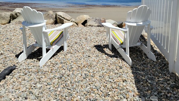 Two white Adirondack chairs with striped cushions on a pebbled beach, overlooking a calm sea with a clear sky.