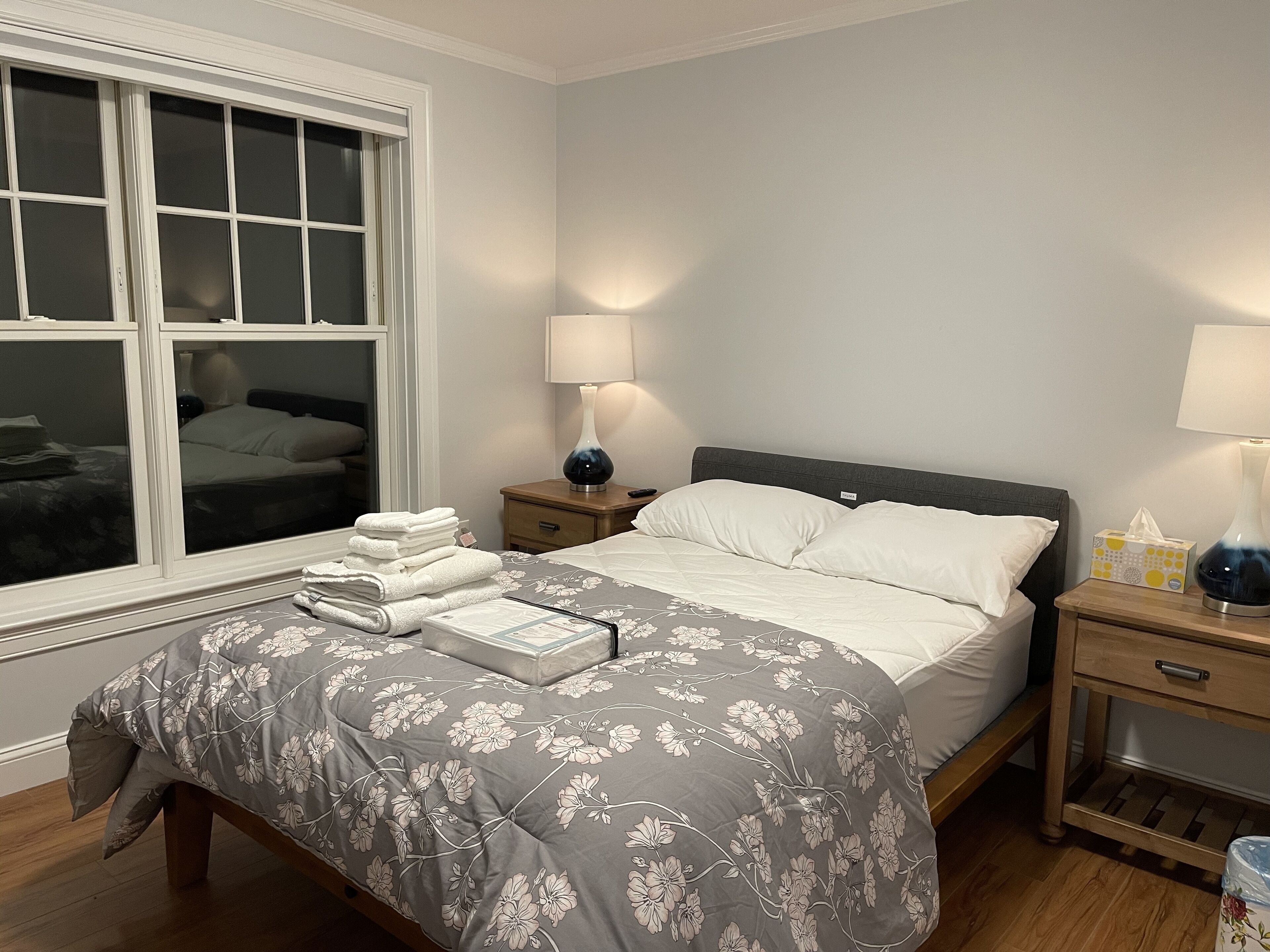 Un lit bien fait, recouvert d’une couvre-lit à motifs floraux, une lampe de chevet et une pile de serviettes pliées sur la table de chevet.