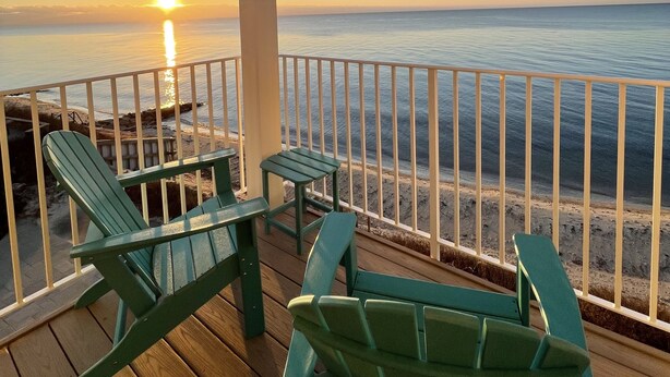 A wooden deck with green Adirondack chairs and a small table, overlooking a beach with a railing. - Cape Cod Oceanfront New Condo with Ocean View & Private Beach, OceanBeachVista! (Dennis)