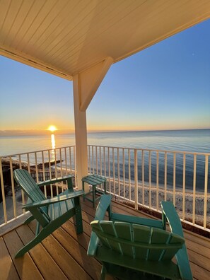 A wooden deck with green Adirondack chairs and a small table, overlooking a beach with a railing.