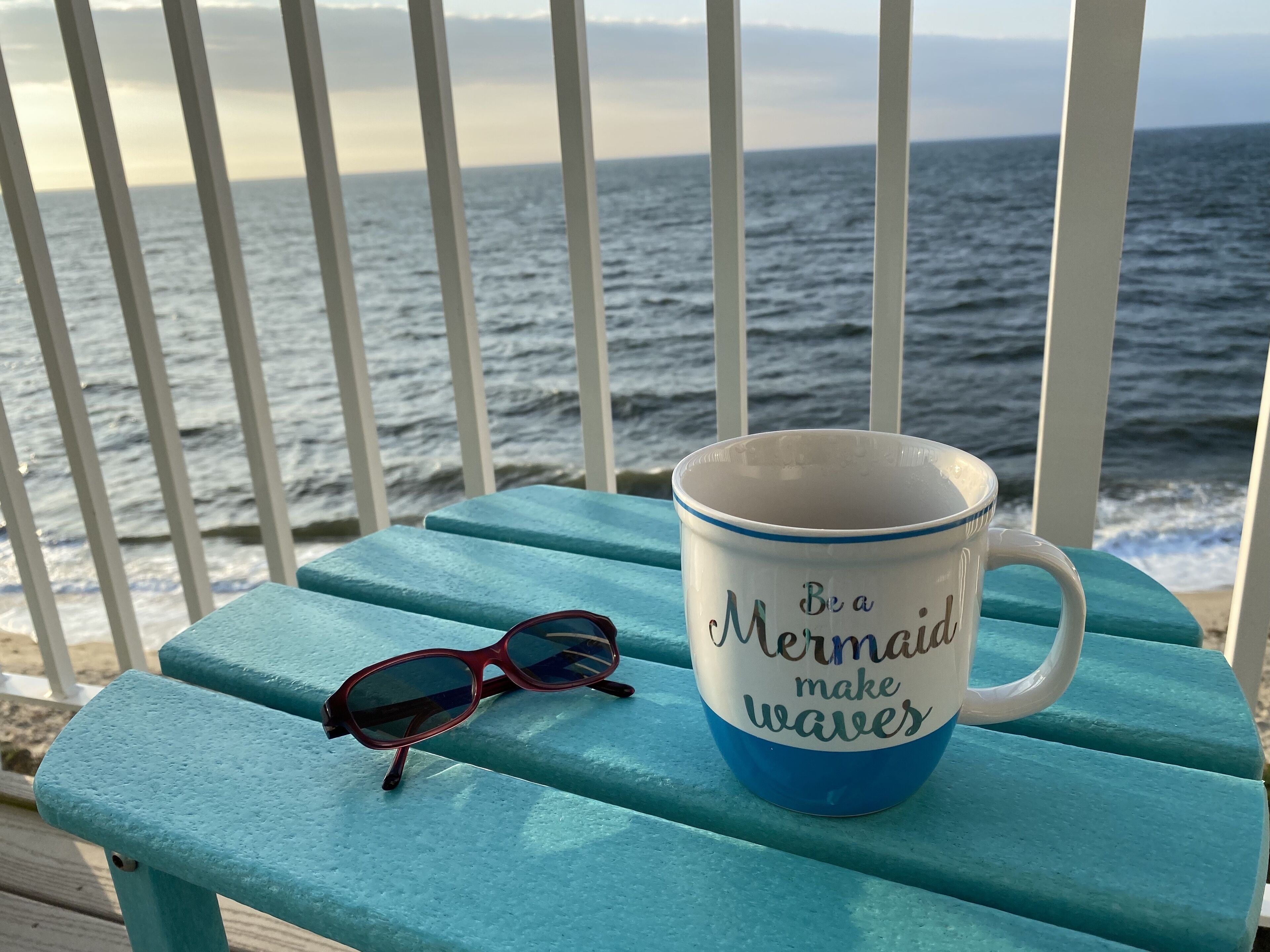 Une tasse ornée d’un motif de sirène et des lunettes de soleil posées sur un banc turquoise, au bord de l’océan.