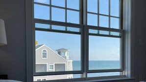 A bedroom with a large window overlooking a beach and a house. - Cape Cod Oceanfront New Condo with Ocean View & Private Beach, OceanBeachVista! (Dennis)