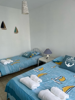 A hotel room with two beds, each with matching blue bedding featuring nautical designs. There are white towels on the beds, a small white table with a lamp, and decorative wall art.