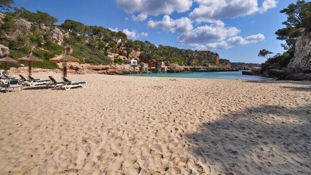 Una spiaggia sabbiosa con lettini e ombrelloni, una scogliera rocciosa sullo sfondo e un cielo azzurro limpido.