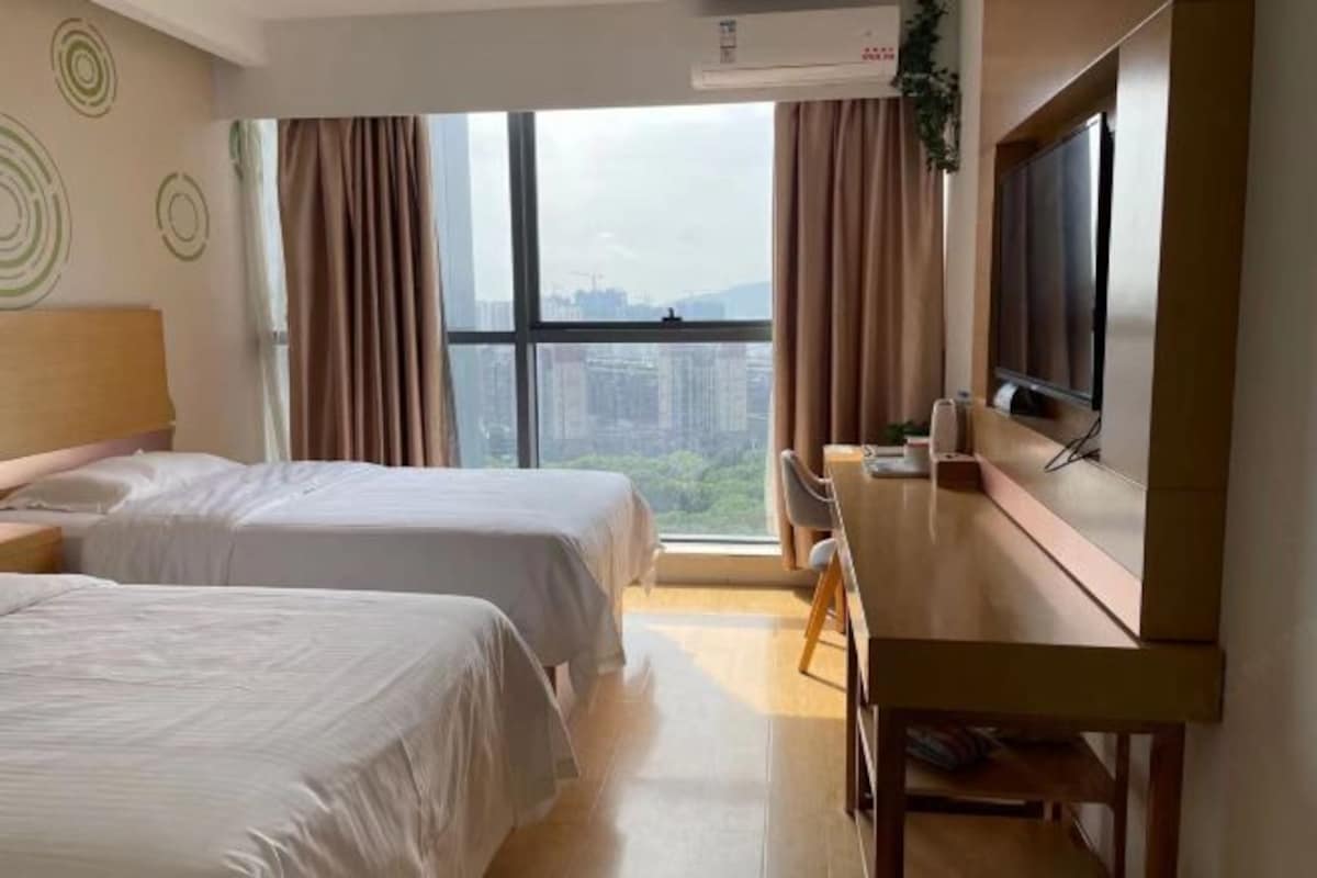 Habitación de hotel con dos camas, un escritorio, un televisor y un amplio ventanal con vistas a la ciudad.
