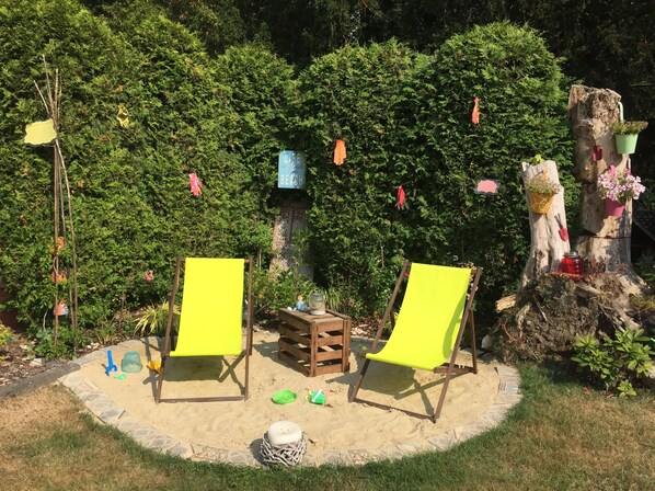 Two yellow chairs in a garden with a sandy area, surrounded by greenery and a tree with hanging decorations.