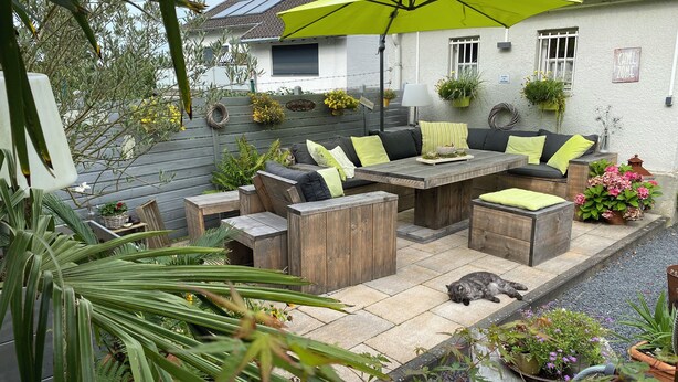 Eine Terrasse mit Holzmöbeln, einem leuchtend gelben Sonnenschirm und Topfpflanzen.