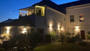 Ein zweistöckiges Haus mit Balkon, beleuchtet mit Lichterketten, umgeben von einem Garten mit Topfpflanzen.