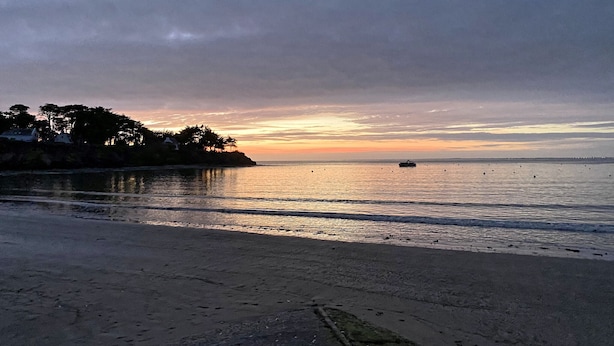 Una playa con mar tranquilo, un barco a lo lejos y un atardecer.