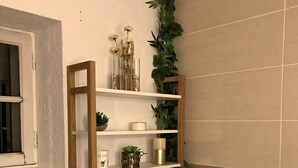 Un baño con un estante blanco, una planta colgante, una cesta de mimbre con una planta y suelo de madera.