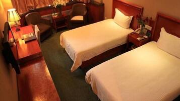 Quarto de hotel com duas camas de solteiro, uma escrivaninha com cadeira, uma luminária e uma pequena mesa com um vaso de flores.