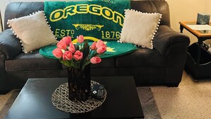 A living room with a couch, a coffee table with a vase of flowers, and a lamp. - Emerald City Cozy Studio Suite - Stay near university fun – 5 min drive (Eugene)