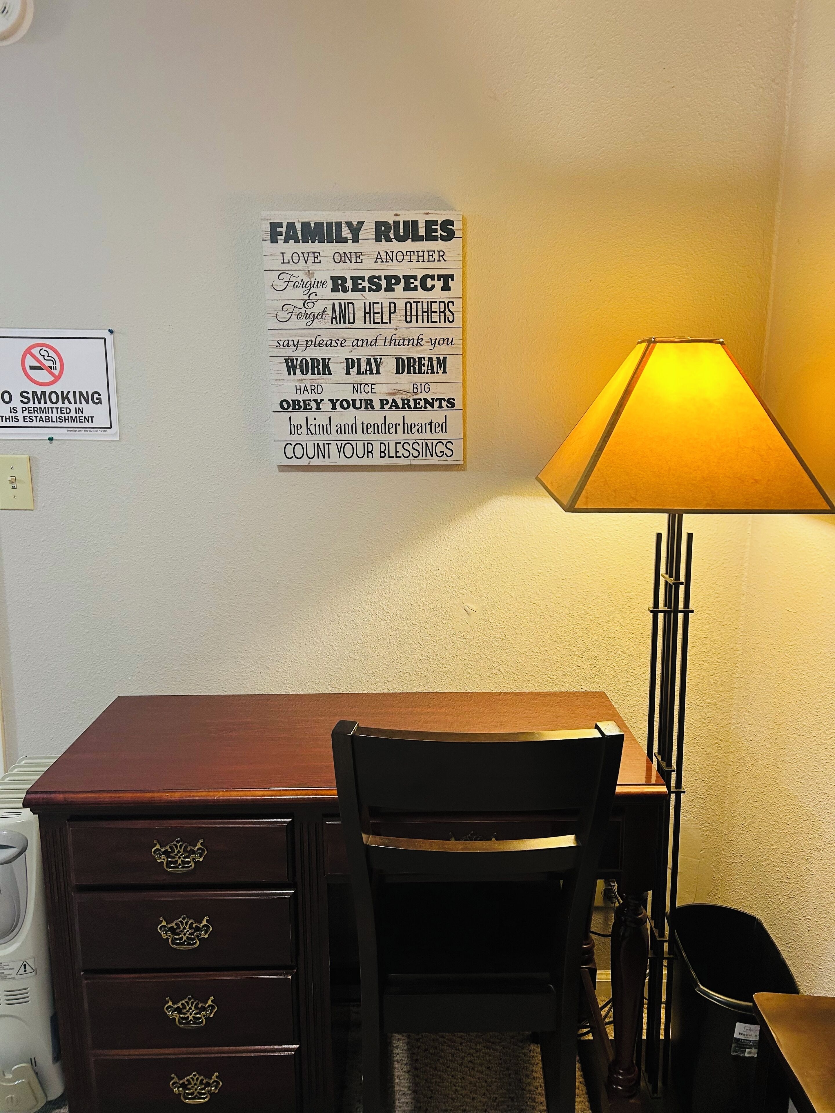 A room with a wooden desk, a chair, a lamp, and a wall with family rules and a no-smoking sign.