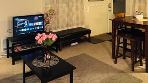 A living room with a television, a bench, a table with a vase of flowers, and a dining area with chairs and a table. - Emerald City Cozy Studio Suite - Stay near university fun – 5 min drive (Eugene)