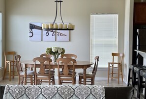 A dining area with a round wooden table, six chairs, and a vase of flowers. There is a chandelier above the table and a window with blinds in the background.