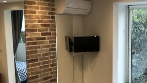 A modern living room with a brick accent wall, a mounted flat-screen TV, and a large sliding glass door leading to a balcony. - Studio Atypique (Fréjus)
